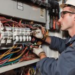 Electrician working on an electrical panel, handling wires and circuit breakers, emphasizing electrical safety and maintenance in fire prevention.